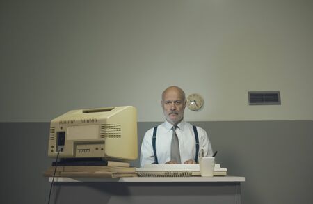 Sad senior businessman working with an outdated computer on a small desk, he is sad and disappointed