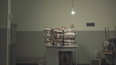 Businessman working behind a lot of stacked paperwork on his desk in a rundown office with cheap furnitureの写真素材