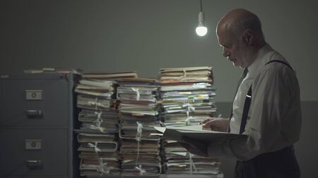 Serious pensive businessman checking a file in a vintage style office and piles of paperwork,...
