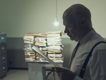 Serious pensive businessman checking a file and piles of paperwork in the background, business and...