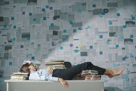 Exhausted business executive lying down on the desk and sleeping, wall covered with financial reports in the background, overtime work and deadlines conceptの写真素材