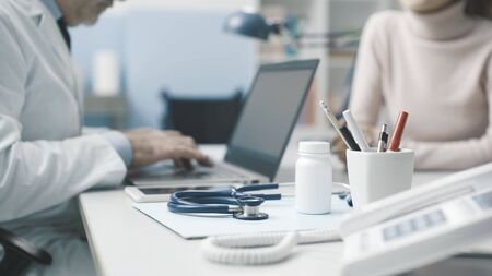 Professional doctor sitting at desk, he is giving a consultation to a patient and typing on a laptopの写真素材