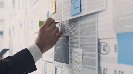 Businessman analyzing a wall with many financial charts and reports, he is underlining text with a...