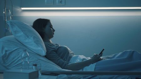 Sleepless worried patient lying in bed late at night at the hospital and chatting with her smartphoneの写真素材
