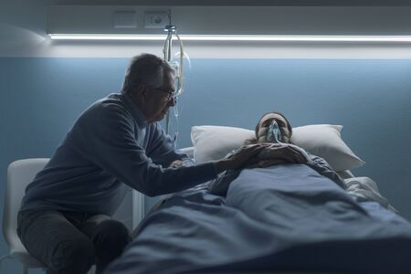 Concerned senior man assisting her young daughter lying in bed at the hospital, he is holding her hand and sitting next to herの写真素材