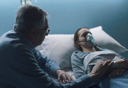 Concerned senior man assisting her young daughter lying in bed at the hospital, he is holding her hand and sitting next to herの写真素材