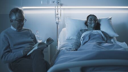 Senior man sitting next to her daughter at the hospital and assisting her at night, he is reading a...
