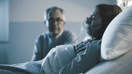 Senior man sitting next to her daughter at the hospital and assisting her at night, she is sleeping...