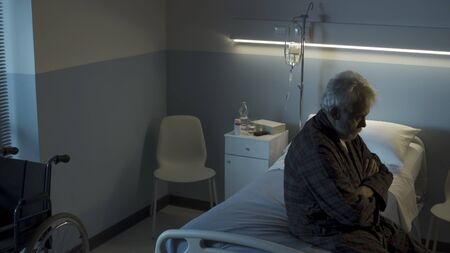 Depressed senior man sitting on the hospital bed alone at night, he feels lonely and sadの写真素材