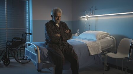 Depressed senior man sitting on the hospital bed alone at night, he feels lonely and abandoned