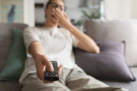 Woman relaxing on the sofa, holding the remote control and watching TV, she is bored and yawning