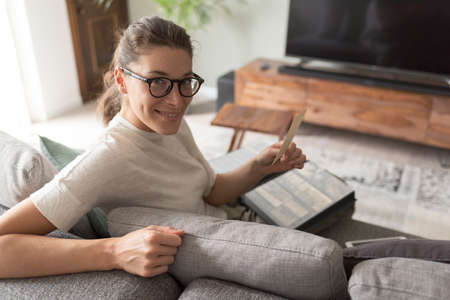 Happy young woman relaxing on the sofa at home and watching pictures in her photo album, lifestyle...
