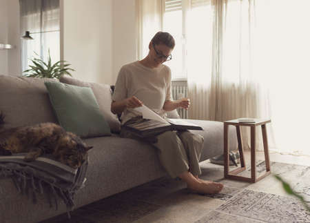 Happy young woman relaxing on the sofa at home and watching pictures in her photo album, lifestyle and hobbies conceptの写真素材