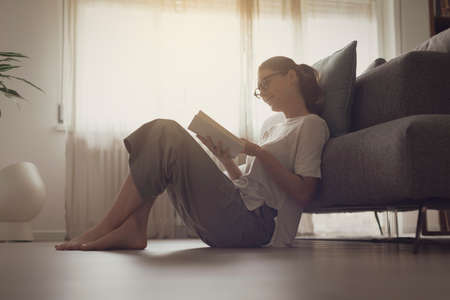 Happy woman with glasses sitting on the floor at home and reading an interesting book