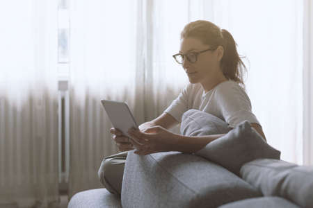 Young attractive woman relaxing at home in the living room, she is sitting on the couch and connecting online with her digital tabletの写真素材