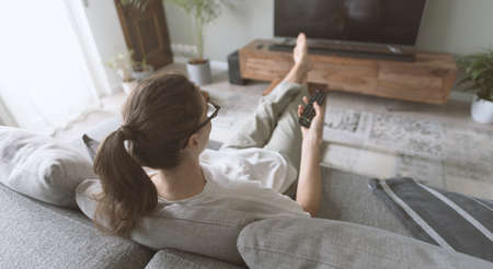 Young woman relaxing on the sofa at home and watching TV, she is holding the remote controlの写真素材