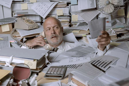 Stressed businessman overwhelmed by work, he is drowning under a lot of paperwork and answering...