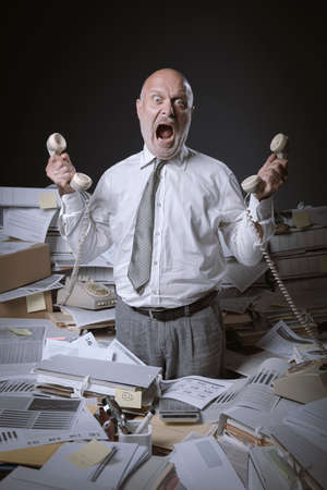Stressed businessman working in a messy office, he is holding many telephone receivers and he is surrounded by lots of paperworkの写真素材