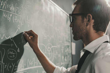 Young smart mathematician drawing math formulas on the chalkboard and thinking: he is solving...