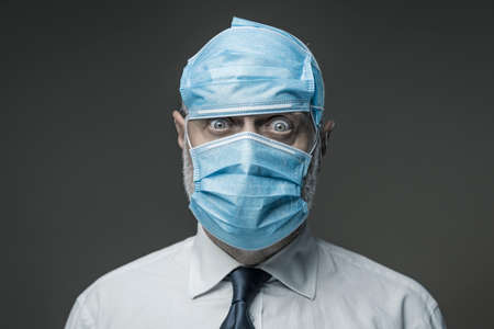 Funny scared businessman wearing many surgical masks on his face and head, coronavirus covid-19 prevention conceptの写真素材