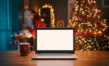 Blank laptop on a desktop at home and Santa Claus bringing gifts in the background, Holidays and Christmas conceptの写真素材