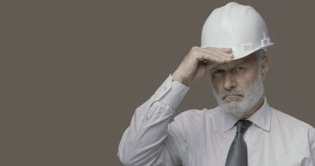 Confident businessman and engineer posing and looking at camera, he is adjusting his safety helmet