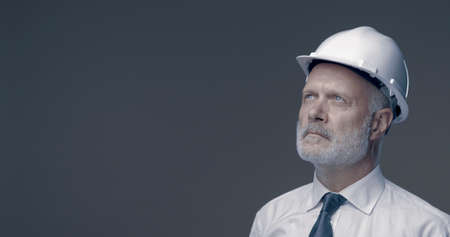 Confident businessman and engineer posing with a safety helmet, he is looking away and thinkingの写真素材