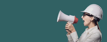 Aggressive businesswoman wearing a safety helmet and shouting through a megaphone, marketing and...