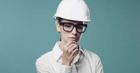 Pensive young businesswoman wearing a safety helmet and thinking with hand on chinの写真素材
