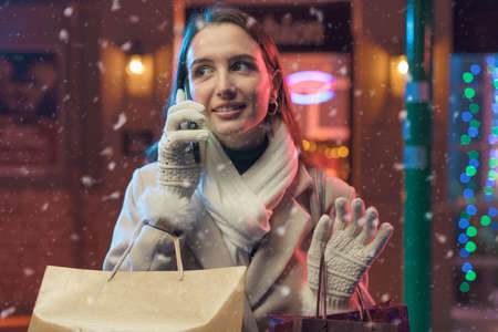 Happy young woman walking in the city streets and doing Christmas shopping, she is carrying many bags and having a phone call with her smartphone
