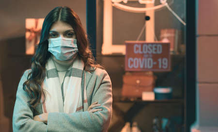 Store closed due to coronavirus covid-19 outbreak, a woman with surgical mask is standing with arms...