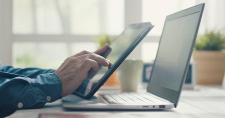 Man sitting at desk and working from home, he is using a digital tablet and a laptopの写真素材