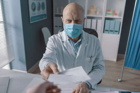 Doctor in his office giving a prescription to the patient, he is wearing a protective surgical mask, point of view shotの写真素材