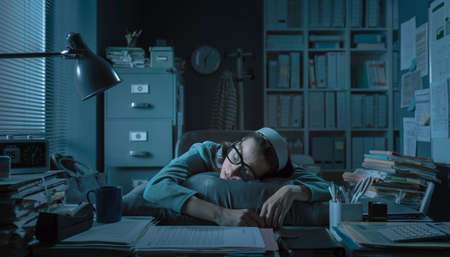 Exhausted young office worker sleeping at her desk, job burnout and overtime work conceptの写真素材