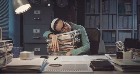 Exhausted office worker sleeping at desk and leaning on a pile of paperwork: overtime work and deadlines conceptの写真素材