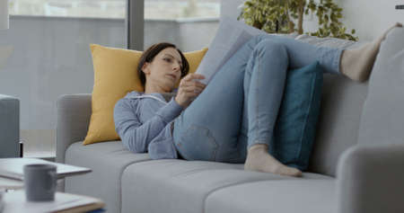 Woman lying on the sofa at home and checking paperwork, remote work conceptの写真素材
