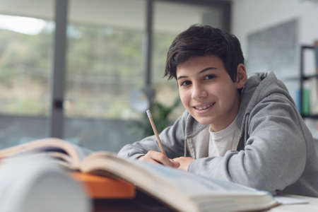 Happy boy sitting at desk and studying at home, he is smiling at camera