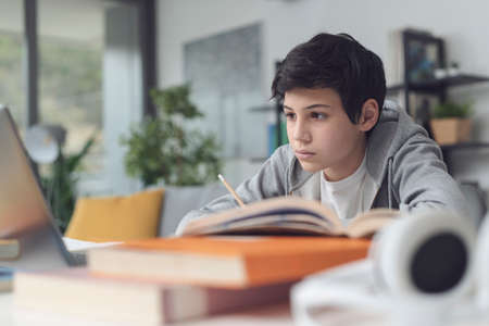 Focused student doing his homework at home, he is writing notes and using a laptopの写真素材
