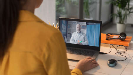 Young woman talking to a doctor online, she is sitting at desk and connecting with her laptop,...