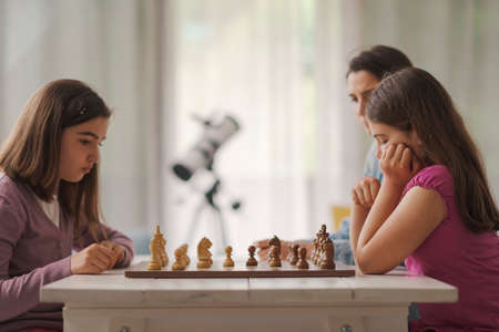 Girls playing chess together at home, their mother is sitting next to them and watchingの写真素材