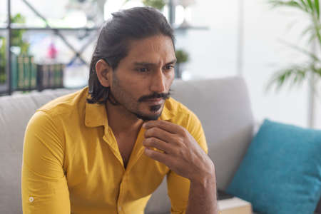 Indian man sitting on the couch at home and thinking with hand on chin