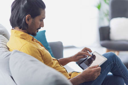 Young confident man sitting on the sofa in the living room and connecting online using a digital...