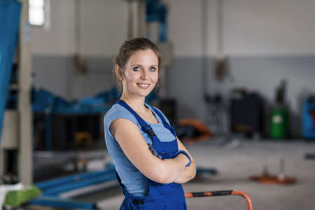 Smiling young female mechanic posing in the car repair shop, she is looking at camera with arms crossedの写真素材