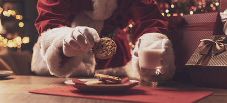 Santa Claus having a delicious snack, he is eating cookies and drinking milk, Christmas and holidays conceptの写真素材