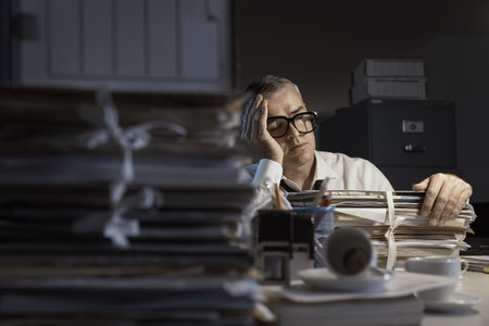 Exhausted businessman sleeping at his desk and working overtime late at night, he is surrounded by piles of paperworkの写真素材