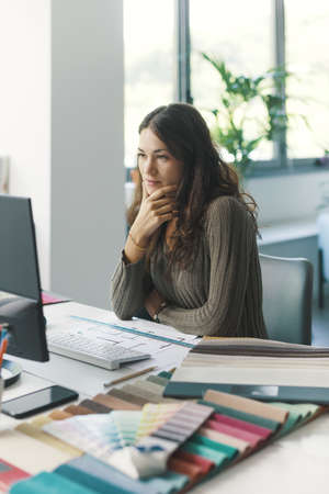 Young professional designer sitting at desk and working with her computer, she is looking at the screen and thinking with hand on chin, ideas and creativity conceptの写真素材