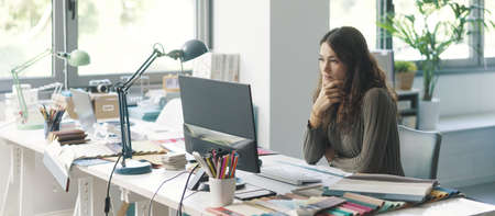 Young professional designer sitting at desk and working with her computer, she is looking at the...