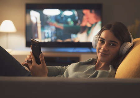 Young woman lying on the couch, she is connecting with her smartphone and looking at camera