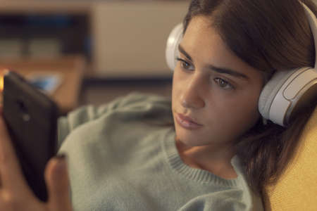 Young woman lying down on the couch and relaxing, she is wearing headphones and connecting with her...
