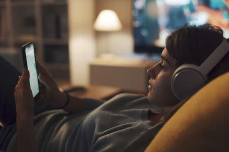 Teenager relaxing on the sofa in the living room, she is wearing headphones and connecting online...